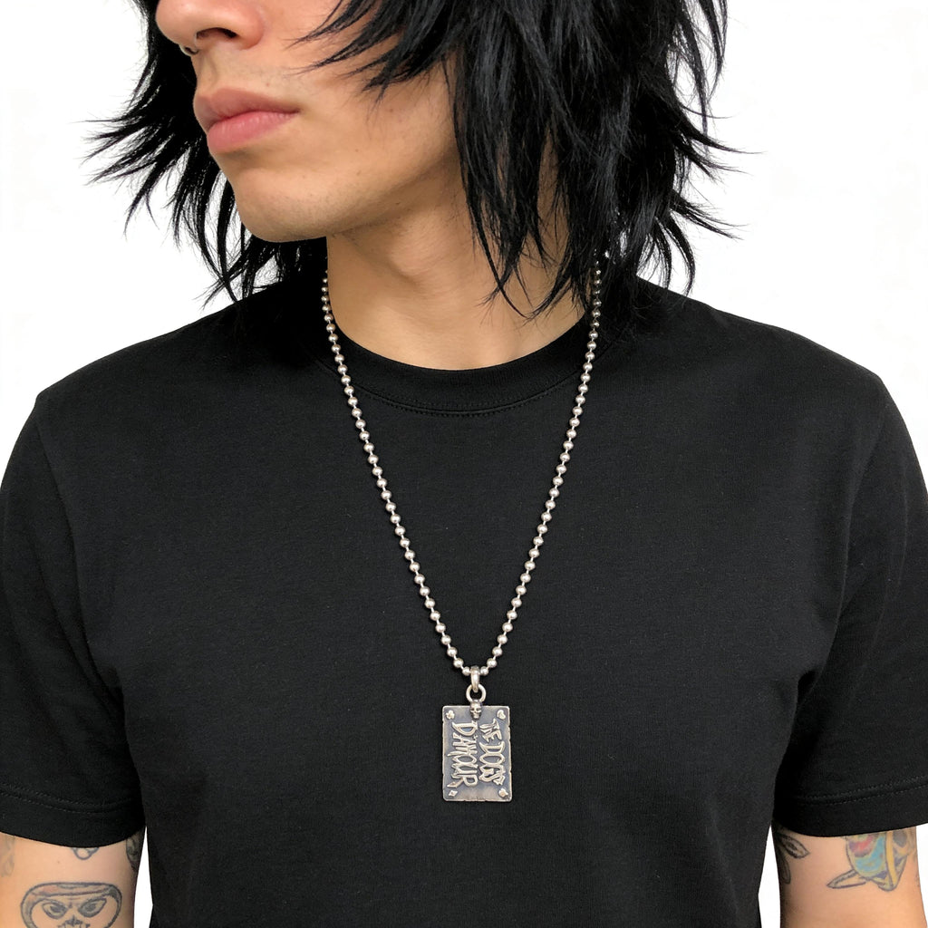 Person wearing a black t-shirt with a silver necklace featuring a rectangular dogs damour pendant.